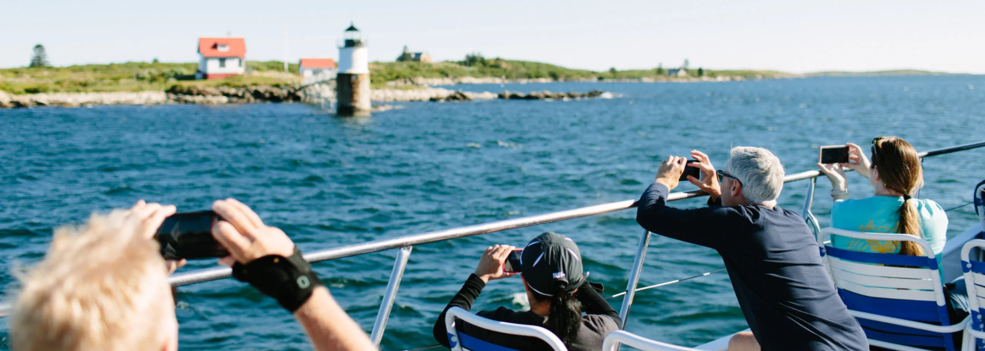 People on a boat taking photos of a distant lighthouse on a sunny day.